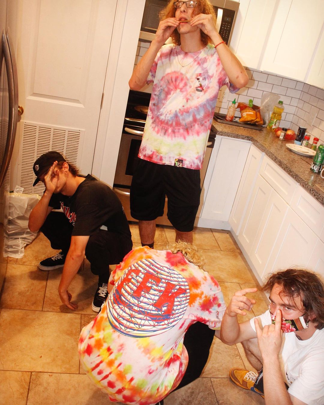 Three people in a kitchen with one person in a tie-dye shirt and another wearing a colorful 'FFK' T-Shirt