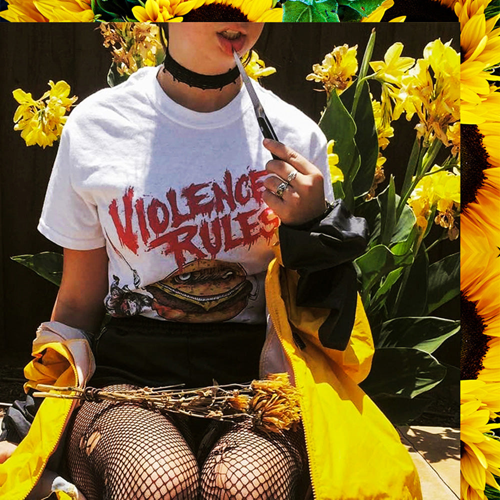 Person wearing a white FFK t-shirt with graphic design and Violence Rules text, surrounded by yellow flowers.